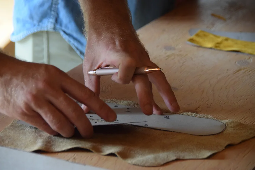 Manos marcando con un punzón el contorno de un patrón de papel sobre una piel curtida al vegetal, apoyada en un tablero de madera